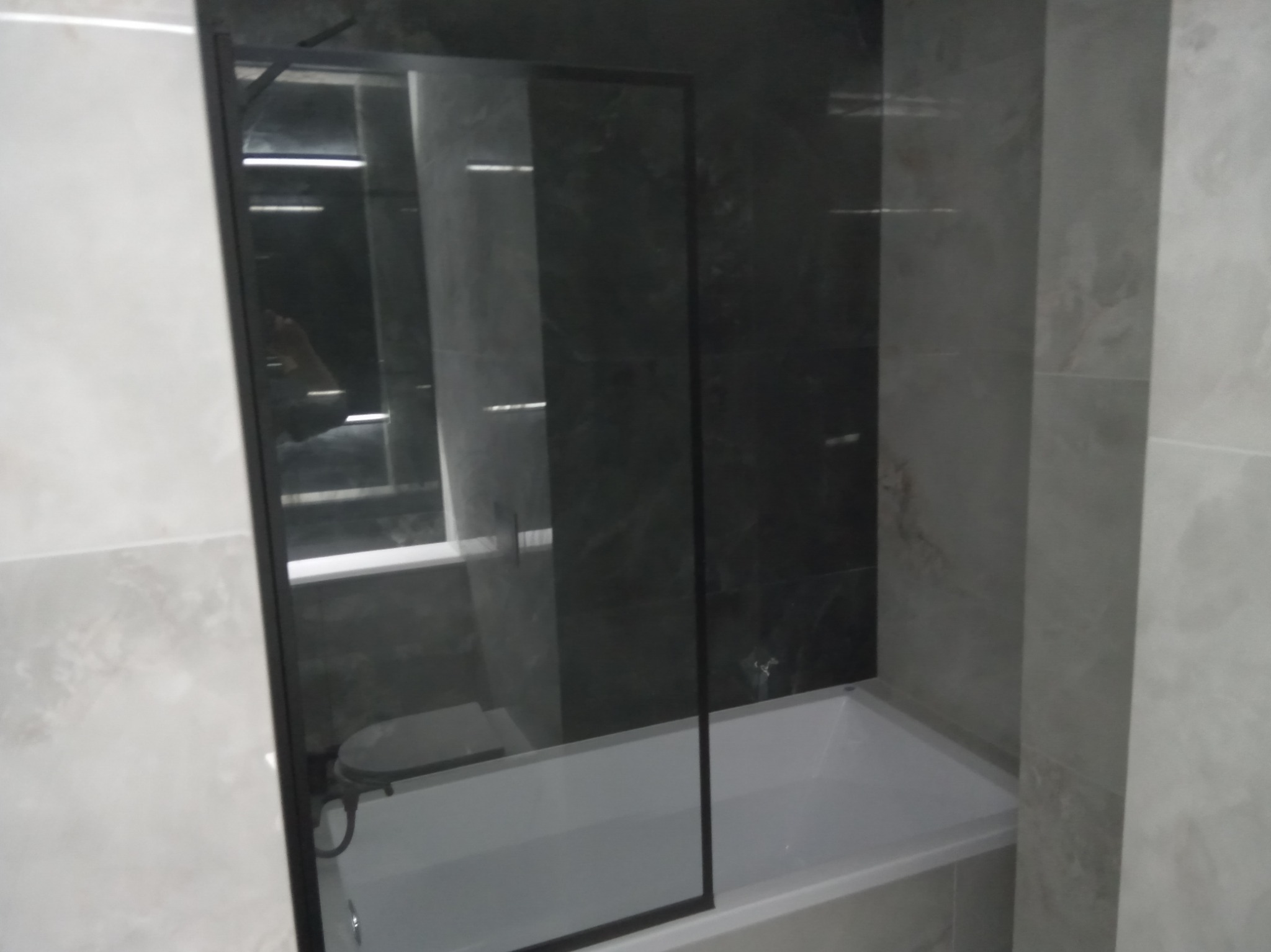 Nowoczesna kabina nawannowa ze szkła hartowanego i czarnymi profilami, zamontowana w wykończonej łazience z szarymi płytkami imitującymi marmur.