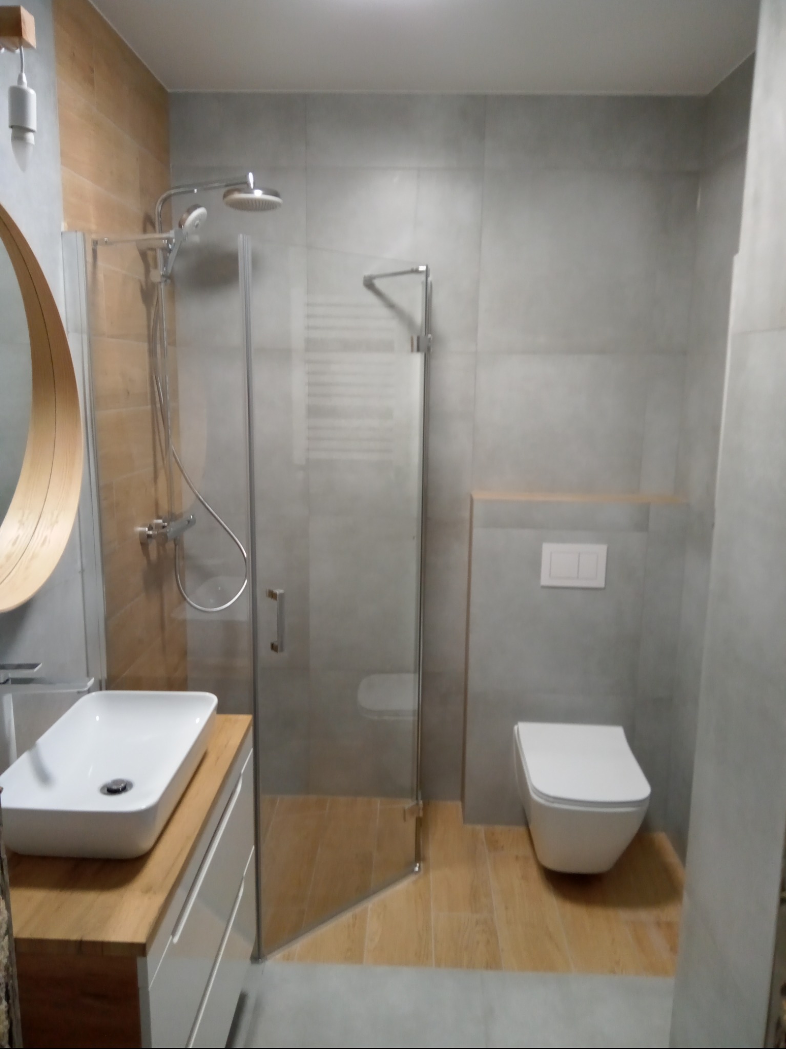 Nowoczesna łazienka z kabiną prysznicową typu walk-in, umywalką nablatową na drewnianym blacie i podwieszaną toaletą. Szare płytki imitujące beton i drewnopodobna podłoga.