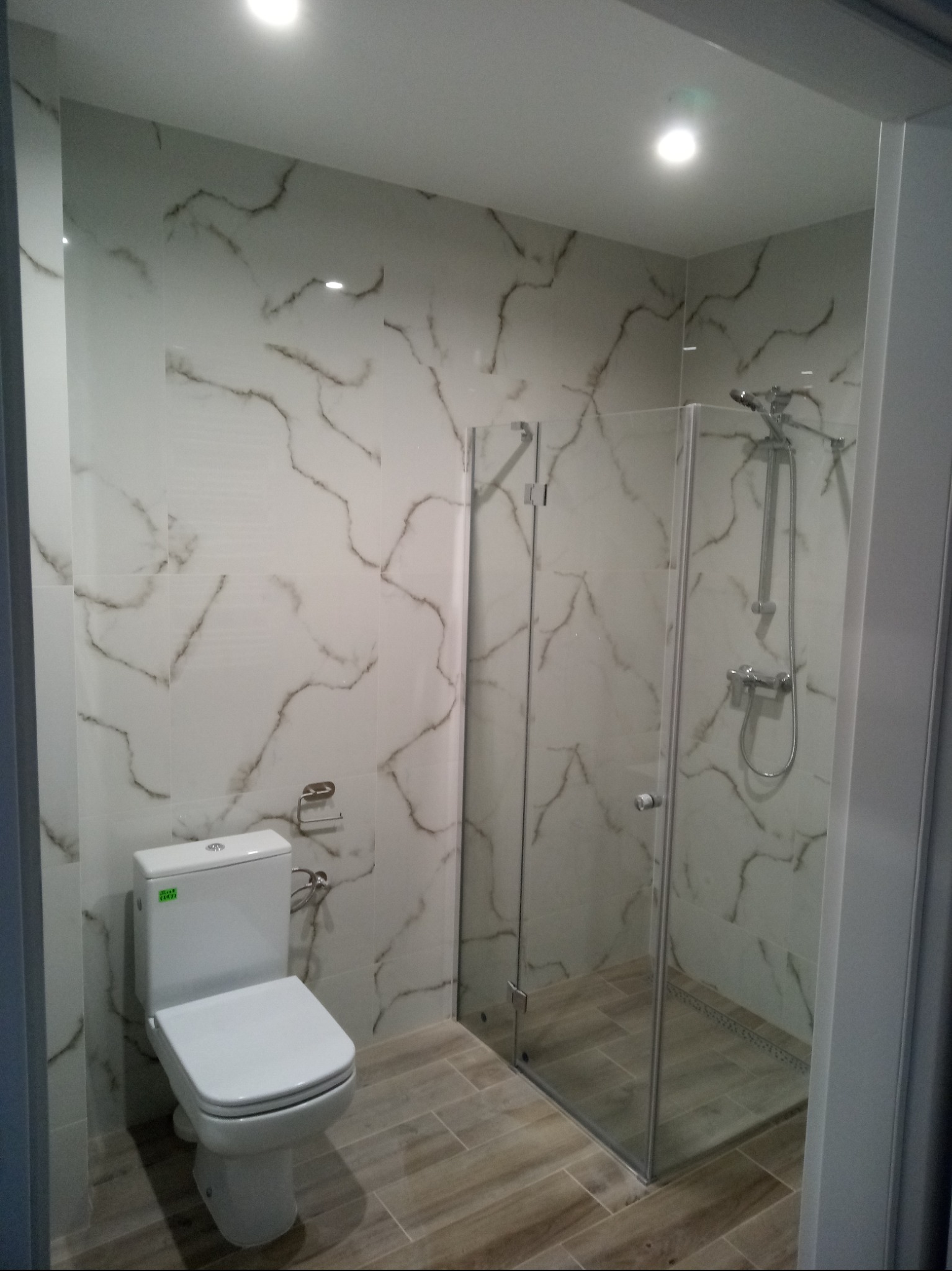 Wykończona łazienka z kabiną prysznicową ze szklanymi drzwiami, toaletą i płytkami imitującymi marmur na ścianach oraz drewnopodobnymi na podłodze.