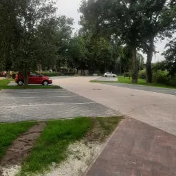 Szeroki kadr na parking z kostki brukowej w różnych kolorach i wzorach, z zaparkowanymi samochodami i drzewami w tle, widoczne ślady opon na trawie.