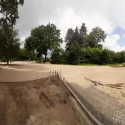 Teren przygotowany pod budowę chodnika z widocznymi krawężnikami, stertami piasku i wbitymi prętami w ziemię, w tle drzewa i budynek.