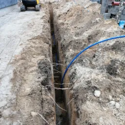 Wąski wykop w ziemi z ułożonymi niebieskimi rurami, widoczny żółty mini-koparka Komatsu w tle, obok budynku w trakcie budowy, zwały ziemi i materiały budowlane w otoczeniu drzew.