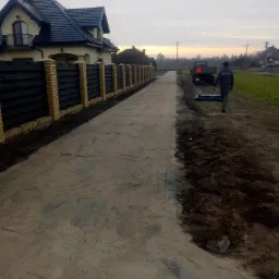 Przygotowanie podłoża pod drogę dojazdową z widocznym ubitym piaskiem, granicą z ziemią, ogrodzeniem z ceglanymi słupkami i drewnianymi panelami, oraz ciężarówką w tle.