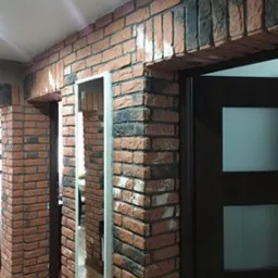 Ściana wykończona dekoracyjną cegłą w odcieniach czerwieni i czerni, z widocznymi futrynami drzwiowymi i fragmentem podłogi z paneli.