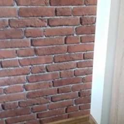 Ściana z tapetą imitującą czerwoną cegłę, widoczna listwa przypodłogowa w kolorze drewna i fragment białej framugi drzwi.