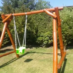 Drewniana huśtawka ogrodowa z zielonym siedziskiem zawieszona na białych linach, widoczna na tle trawnika i zieleni ogrodowej.