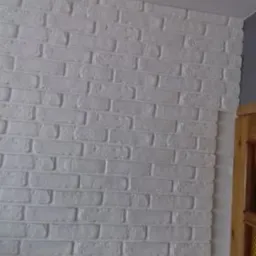 Ściana wykończona białymi, dekoracyjnymi płytkami imitującymi cegłę, widoczny fragment futryny drzwi i pomalowany na szaro narożnik sufitu.