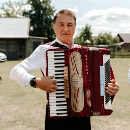 AMIGO ZESPÓŁ MUZYCZNY NOWA SARZYNA - Mężczyzna w białej koszuli gra na akordeonie Hohner Verdi V na tle zielonego trawnika i zabudowań. Instrument w kolorze czerwonym z dekoracyjnymi elementami.