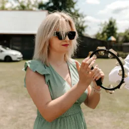 Kobieta w zielonej sukience gra na tamburynie na zewnątrz, w tle budynek i samochód. Letnie oświetlenie, delikatne kolory.