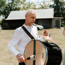 AMIGO ZESPÓŁ MUZYCZNY NOWA SARZYNA - Muzyk z siwymi włosami, w białej koszuli, gra na bębnie basowym z talerzem perkusyjnym na zielonym tle. Widoczny budynek i samochód.