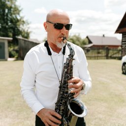 AMIGO ZESPÓŁ MUZYCZNY NOWA SARZYNA - Saksofonista w okularach przeciwsłonecznych gra na saksofonie tenorowym na tle wiejskiego krajobrazu z domem.
