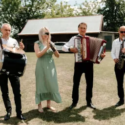 Czteroosobowy zespół muzyczny na trawie przed budynkiem. Perkusista, akordeonista, saksofonista i kobieta z instrumentem perkusyjnym. Białe koszule, czarne spodnie, plener.