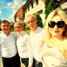 AMIGO ZESPÓŁ MUZYCZNY NOWA SARZYNA - Zespół muzyczny w białych koszulach pozuje do zdjęcia na zewnątrz, z budynkiem w tle i błękitnym niebem z chmurami. Selfie zrobione przez kobietę w okularach.