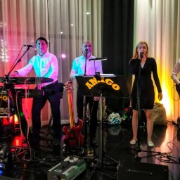 AMIGO ZESPÓŁ MUZYCZNY NOWA SARZYNA - Zespół muzyczny 'Amigo' na scenie: wokalistka w czarnej sukience, klawiszowiec, gitarzysta i saksofonista. Scena oświetlona kolorowymi światłami.