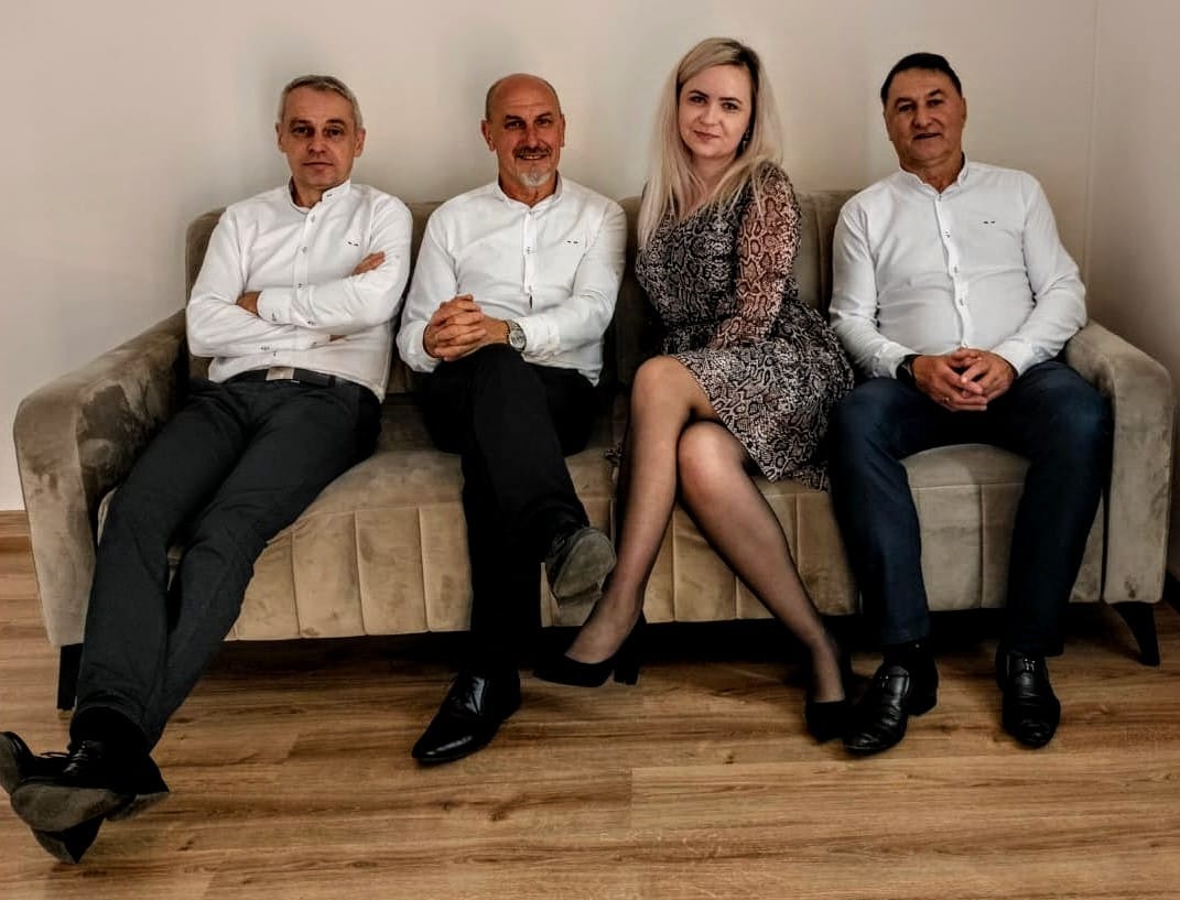 Czteroosobowa grupa, trzech mężczyzn w białych koszulach i ciemnych spodniach oraz blondynka w wzorzystej sukience, siedząca na beżowej kanapie na tle jasnej ściany i podłogi z paneli.