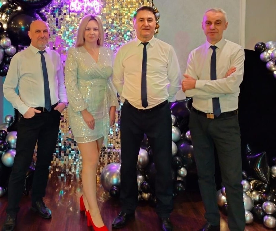 Czteroosobowa grupa w strojach formalnych pozuje na tle błyszczącej ściany i balonów, kobieta w cekinowej sukience i czerwonych butach pośrodku.