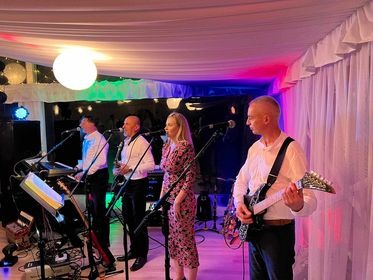 Czteroosobowy zespół grający na scenie: mężczyźni w białych koszulach i czarnych muchach grają na gitarze, keyboardzie i śpiewają do mikrofonu; kobieta w kwiecistej sukience śpiewa do mikrofonu;...