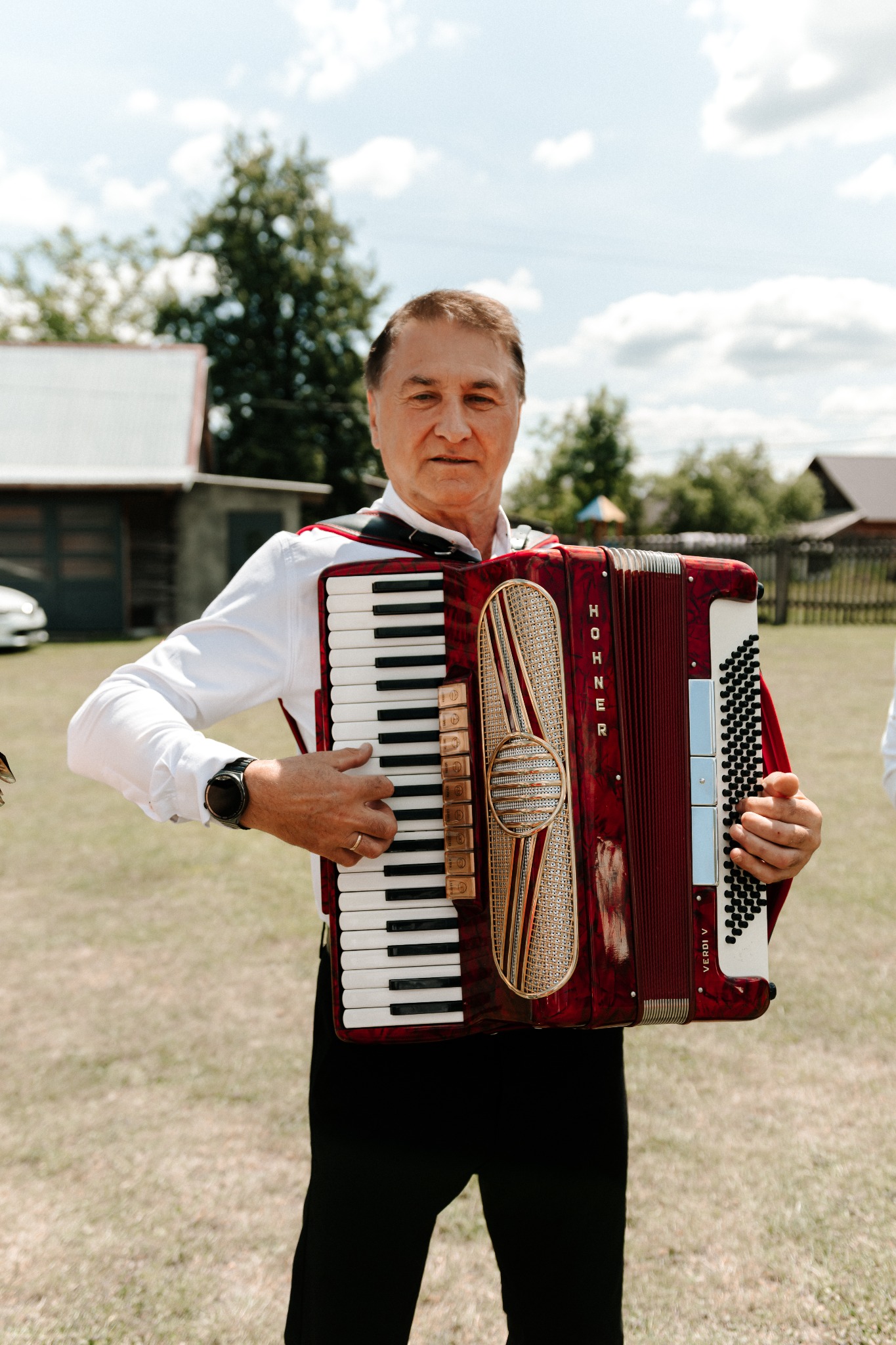 Mężczyzna w białej koszuli gra na akordeonie Hohner Verdi V na tle zielonego trawnika i zabudowań. Instrument w kolorze czerwonym z dekoracyjnymi elementami.