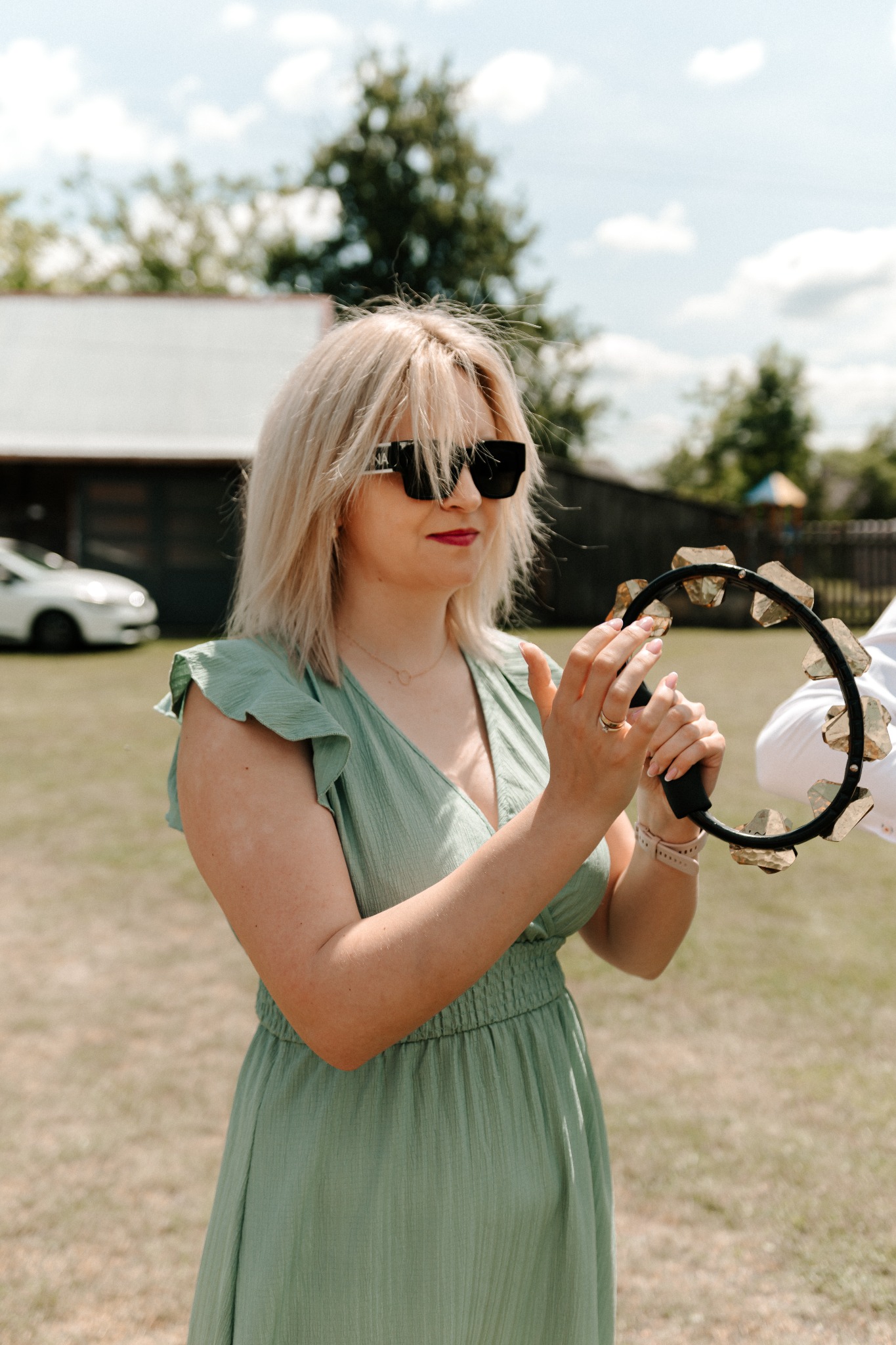 Kobieta w zielonej sukience gra na tamburynie na zewnątrz, w tle budynek i samochód. Letnie oświetlenie, delikatne kolory.