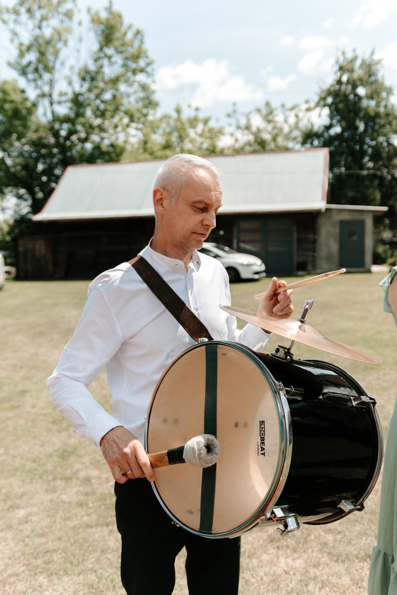 Muzyk z siwymi włosami, w białej koszuli, gra na bębnie basowym z talerzem perkusyjnym na zielonym tle. Widoczny budynek i samochód.