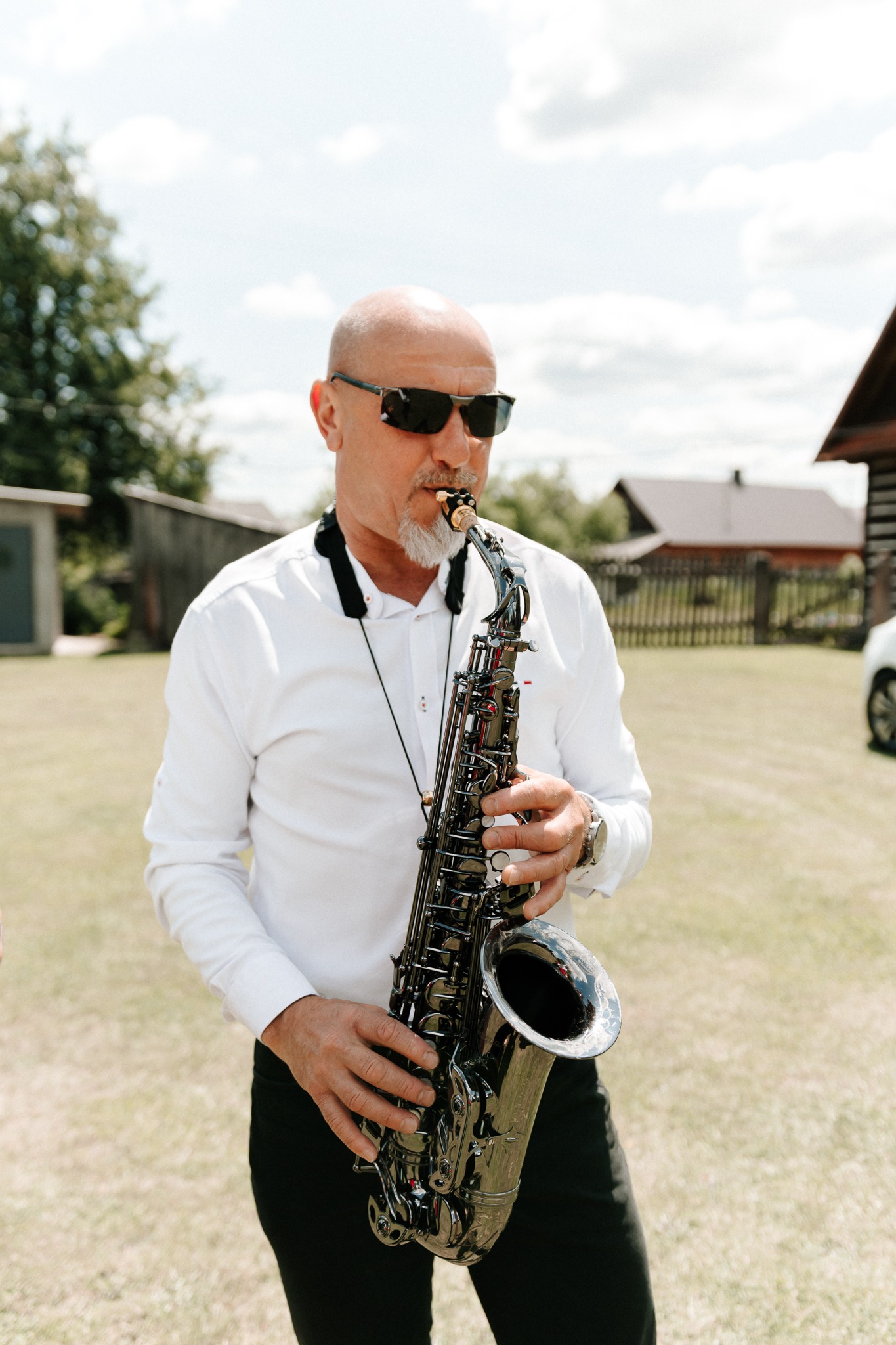 Saksofonista w okularach przeciwsłonecznych gra na saksofonie tenorowym na tle wiejskiego krajobrazu z domem.