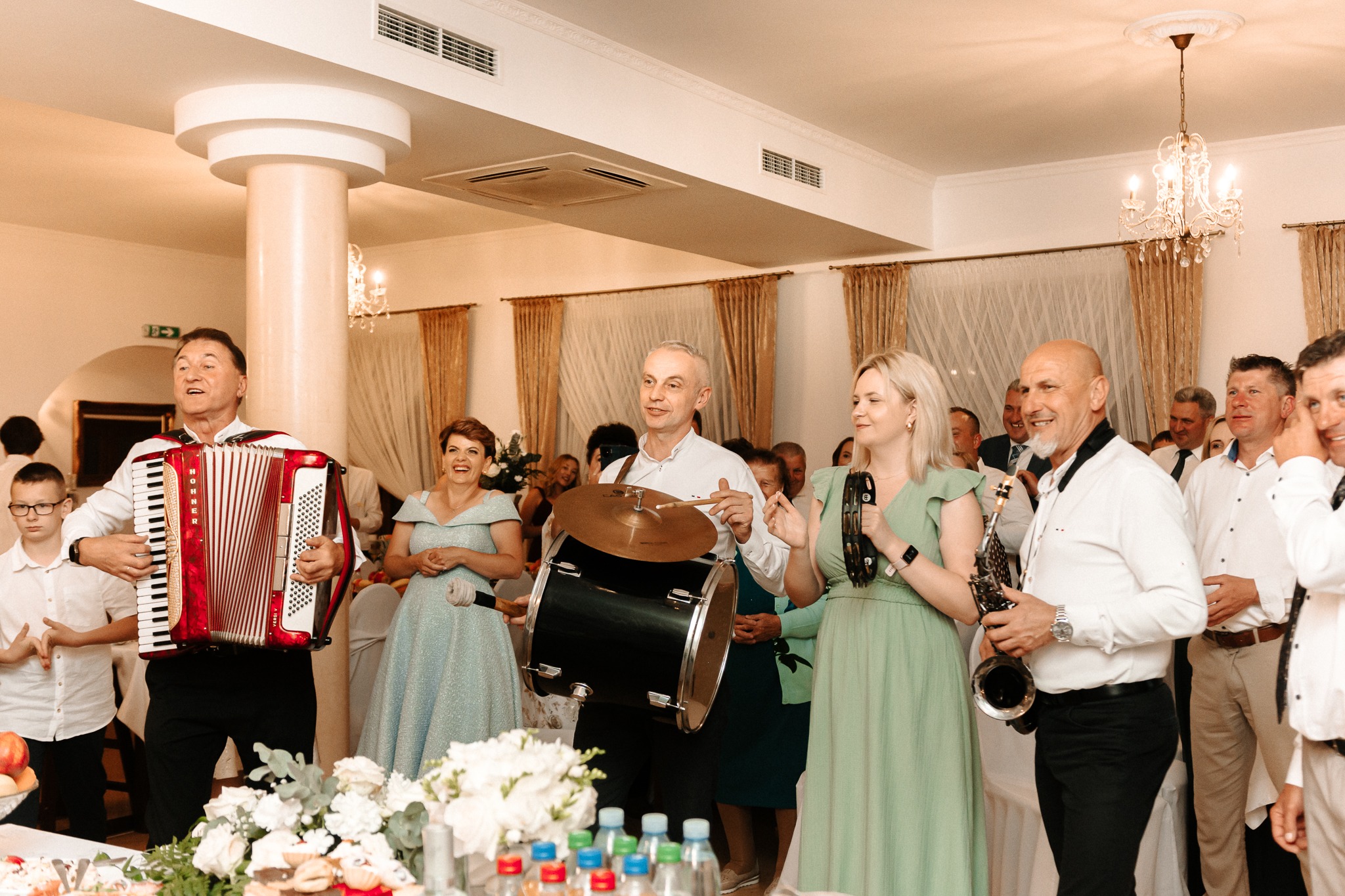 Zespół muzyczny grający na weselu: akordeon, bęben z talerzem, tamburyn i saksofon. W tle goście weselni i dekoracje kwiatowe.