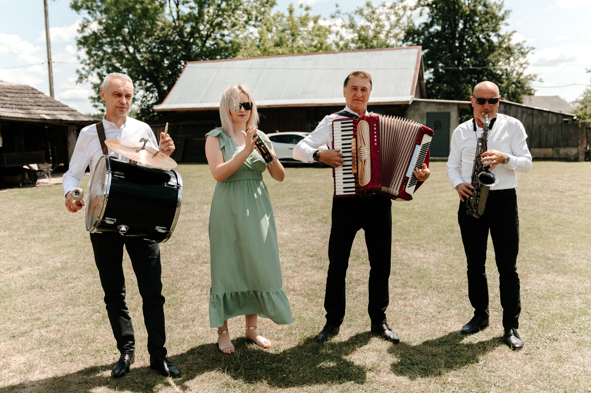 Czteroosobowy zespół muzyczny na trawie przed budynkiem. Perkusista, akordeonista, saksofonista i kobieta z instrumentem perkusyjnym. Białe koszule, czarne spodnie, plener.