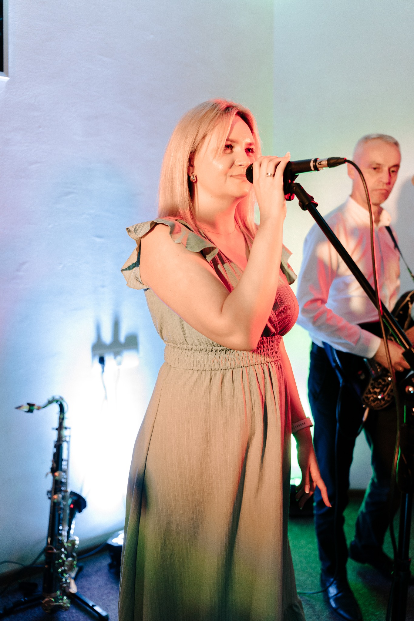 Wokalistka z mikrofonem na scenie, obok saksofon i muzyk z instrumentem. Scena oświetlona kolorowymi światłami, tworzącymi przyjemną atmosferę.