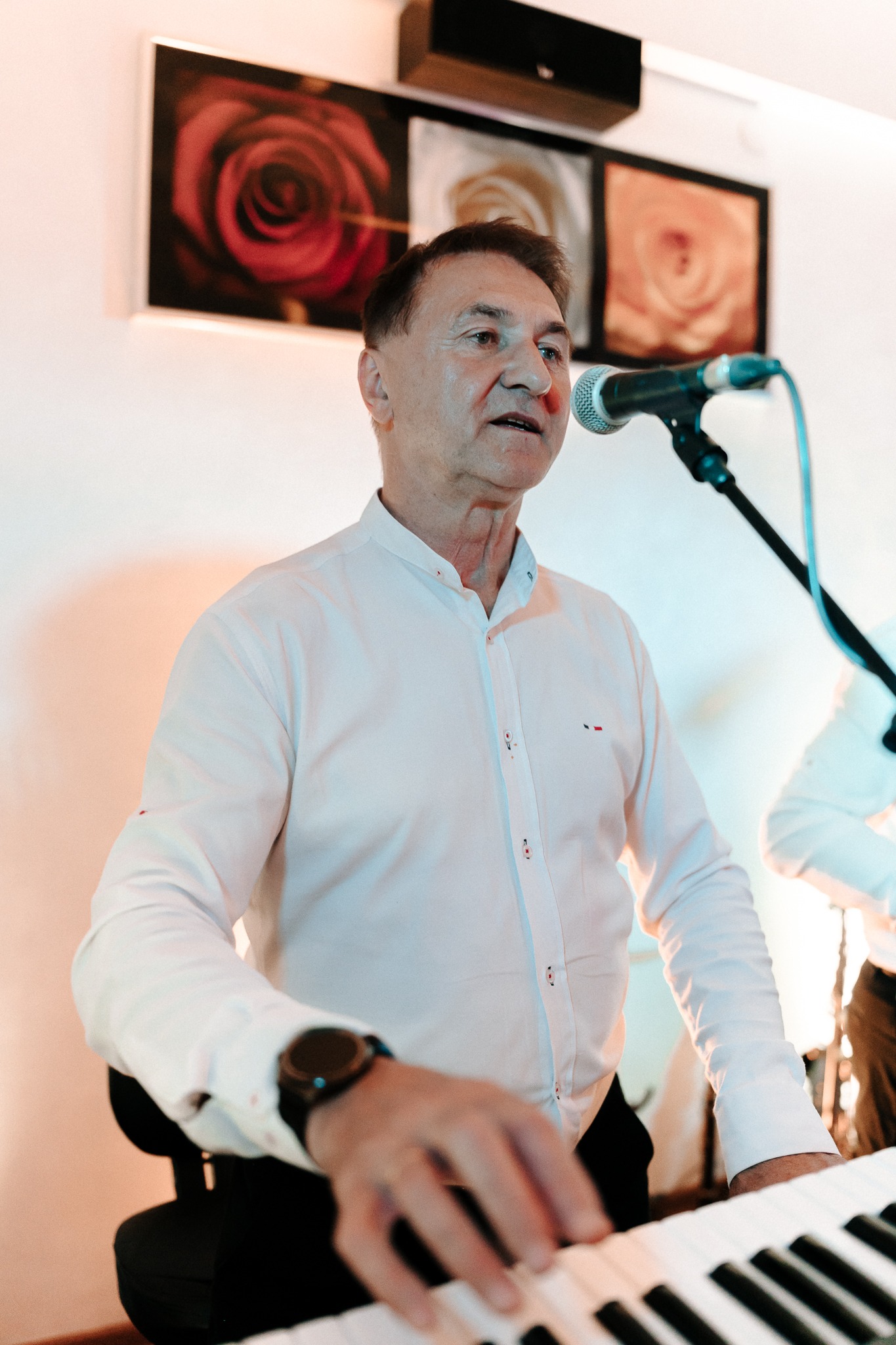 Wokalista grający na keyboardzie, śpiewający do mikrofonu, w tle obrazy z motywem kwiatowym.