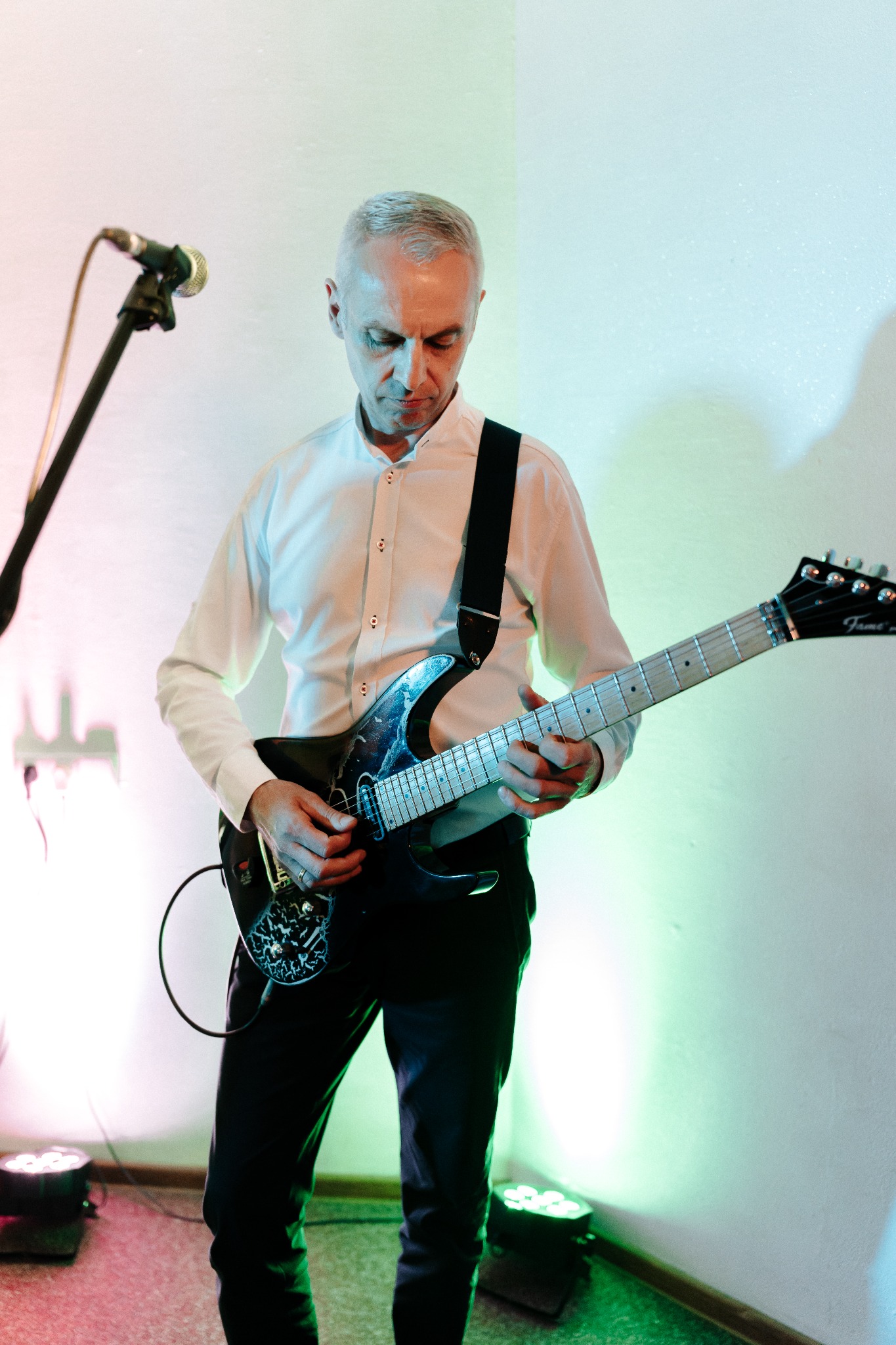 Gitarzysta w białej koszuli gra na czarnej gitarze elektrycznej Fame na tle ściany oświetlonej kolorowymi światłami scenicznymi. Mikrofon w tle.
