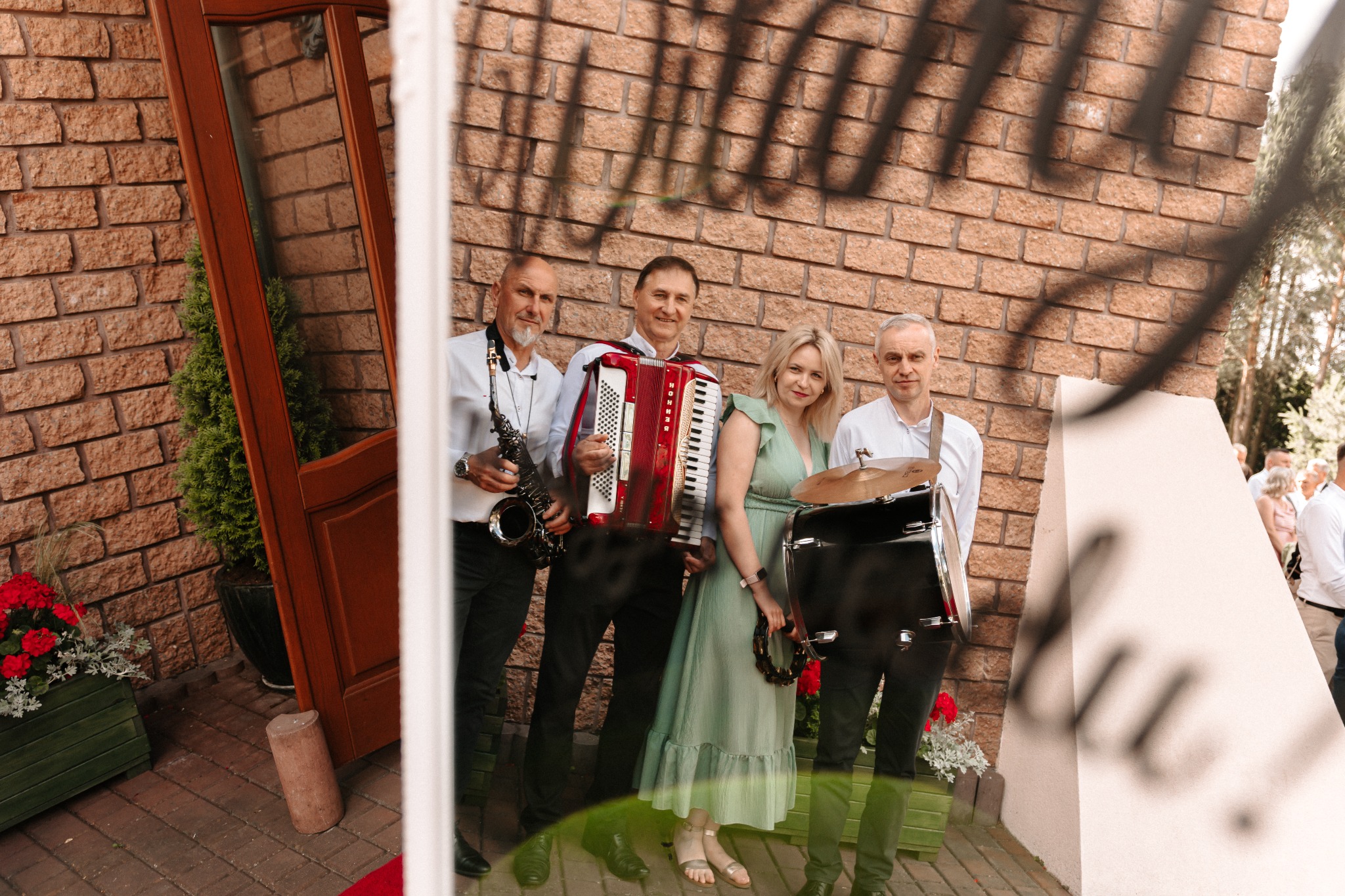 Zespół muzyczny z akordeonem, saksofonem i perkusją pozuje na tle ceglanej ściany, odbity w lustrze. Widoczny fragment czerwonego dywanu i dekoracje kwiatowe.
