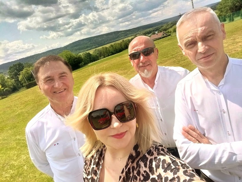 Czteroosobowy zespół muzyczny pozuje do selfie na tle zielonej łąki i wzgórz. Wszyscy uśmiechnięci, ubrani w białe koszule, z wyjątkiem kobiety w panterkowym topie.