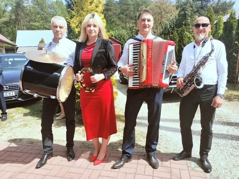 Zespół muzyczny prezentuje się z instrumentami: akordeon, saksofon, bęben i tamburyn. W tle budynek i auto.