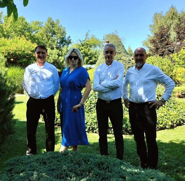 Czteroosobowy zespół muzyczny w eleganckich strojach pozuje w plenerze, otoczony zielenią drzew i krzewów, w słoneczny dzień. Ujęcie od frontu, pełne postacie.