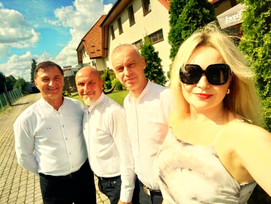 Zespół muzyczny w białych koszulach pozuje do zdjęcia na zewnątrz, z budynkiem w tle i błękitnym niebem z chmurami. Selfie zrobione przez kobietę w okularach.