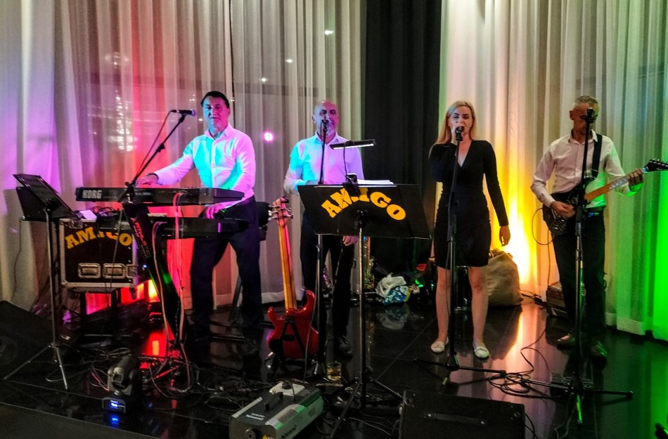 Zespół muzyczny 'Amigo' na scenie: wokalistka w czarnej sukience, klawiszowiec, gitarzysta i saksofonista. Scena oświetlona kolorowymi światłami.