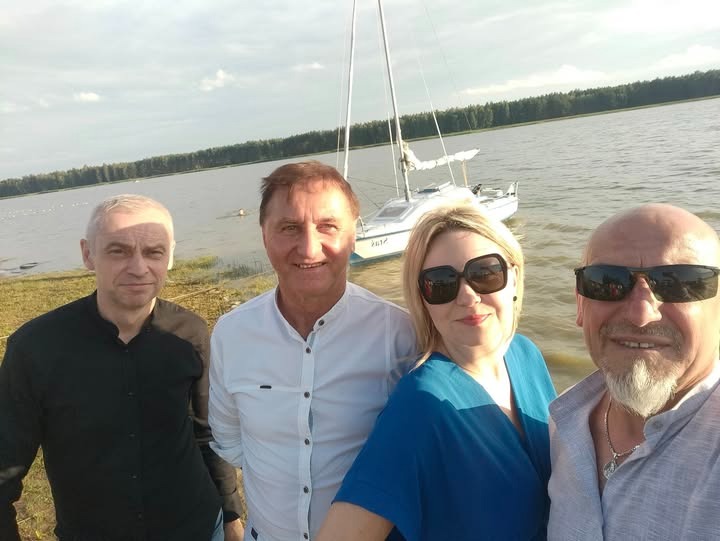 Czteroosobowy zespół muzyczny pozuje na tle jeziora z łódką. Uśmiechnięci muzycy w okularach przeciwsłonecznych i koszulach. Słoneczny dzień nad wodą.