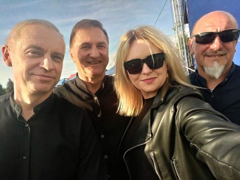 Zdjęcie zespołu muzycznego w czarnych strojach, z blondwłosą wokalistką w skórzanej kurtce i okularach przeciwsłonecznych, robione prawdopodobnie przed występem na tle sceny.