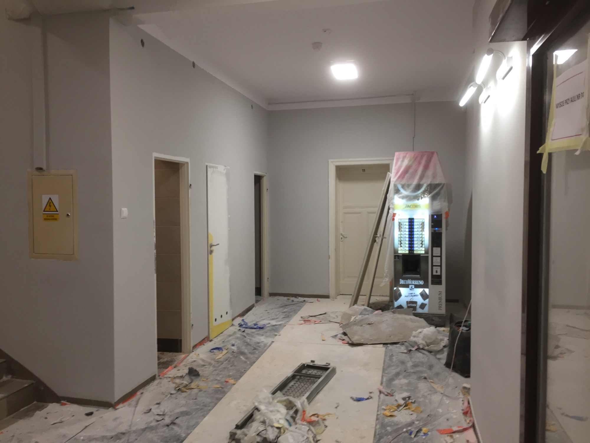 Korytarz w trakcie remontu: szare ściany, zabezpieczona folią podłoga, drabina oparta o ścianę, automat z kawą w folii ochronnej, widoczny rozgardiasz i narzędzia.