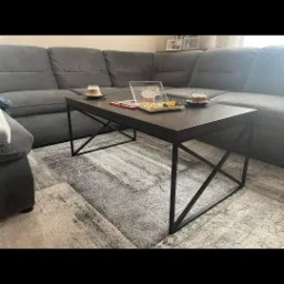 Stolik kawowy z czarnym, matowym blatem i metalową konstrukcją nóg, na blacie filiżanki z kawą, pudełko z czekoladkami i ramka na zdjęcia, ustawiony na wzorzystym dywanie w salonie z szarą sofą.