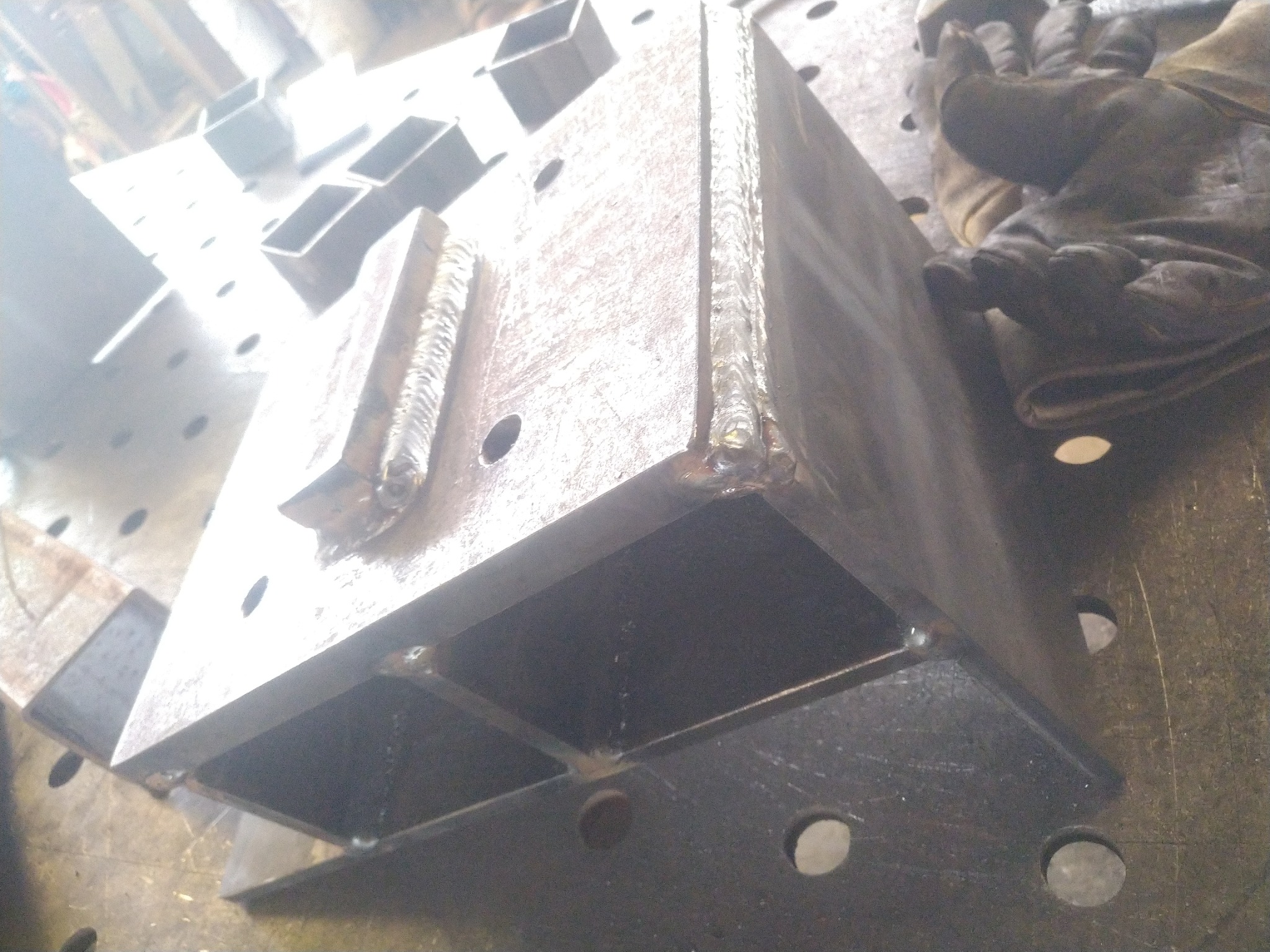 Spawana konstrukcja stalowa z profili zamkniętych, widoczne spoiny i otwory technologiczne na stole spawalniczym, rękawice robocze w tle.