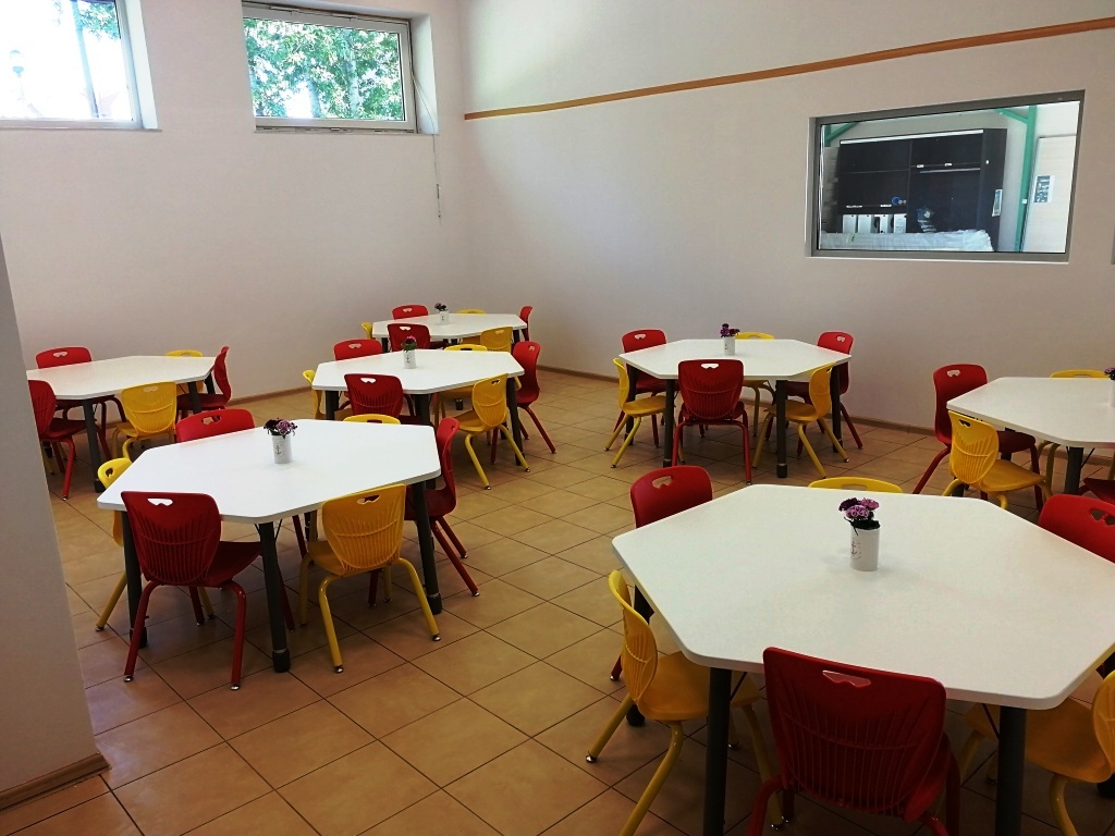 Jasna sala jadalna z sześciokątnymi stołami i krzesłami w kolorach czerwonym i żółtym, małe bukiety kwiatów na stołach, okno z widokiem na zewnątrz.