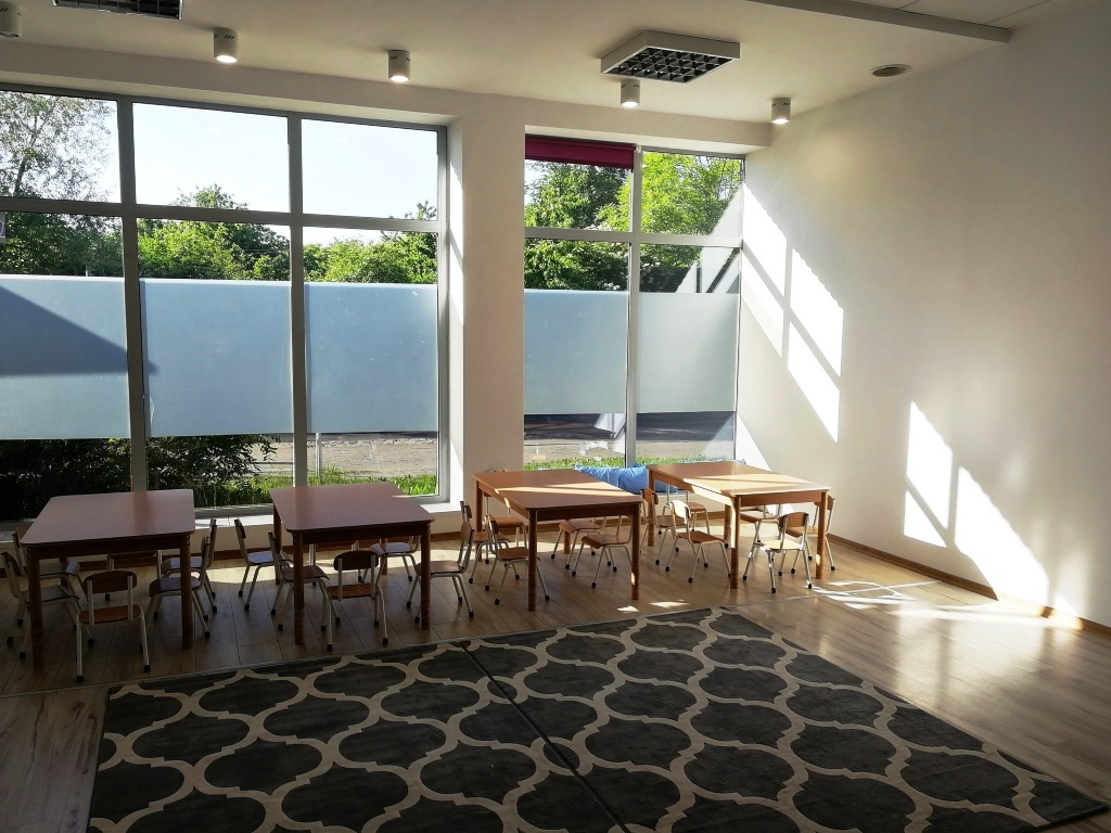 Jasne wnętrze sali z drewnianymi stolikami i krzesłami dla dzieci, duże okna z częściowo matową folią, wzorzysty dywan na podłodze, promienie słoneczne wpadające do środka.