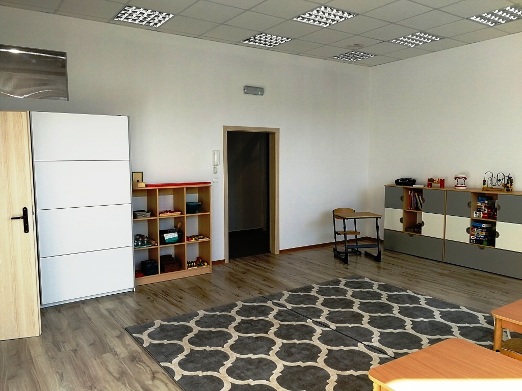 Jasne, przestronne wnętrze sali zabaw z drewnianą podłogą, wzorzystym dywanem, regałami z zabawkami i niskimi stolikami.