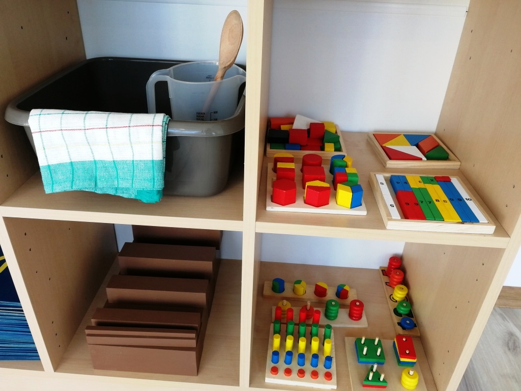 Półka z drewnianymi zabawkami edukacyjnymi: kolorowe klocki, układanki geometryczne, liczydła i naczynia do zabaw sensorycznych.