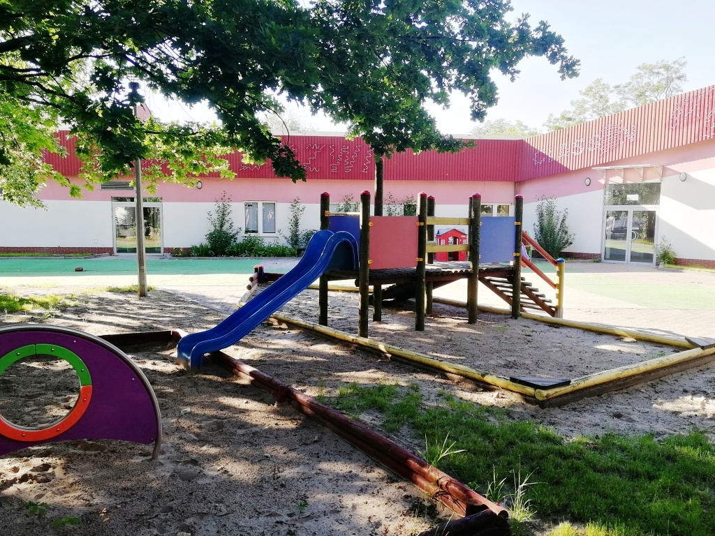 Plac zabaw żłobka z niebieską zjeżdżalnią, drabinkami i piaskownicą na piaszczystym podłożu, na tle budynku z czerwonym dachem i dekoracjami na ścianie.