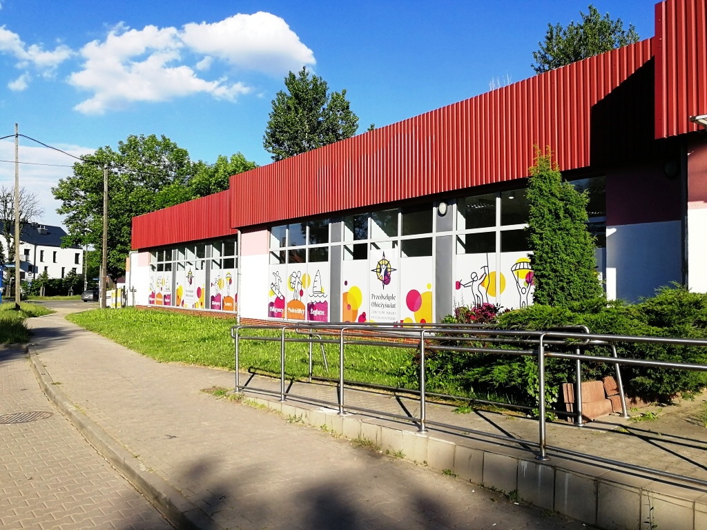 Budynek przedszkola z czerwonymi elementami elewacji i kolorowymi grafikami na oknach, widok z chodnika.