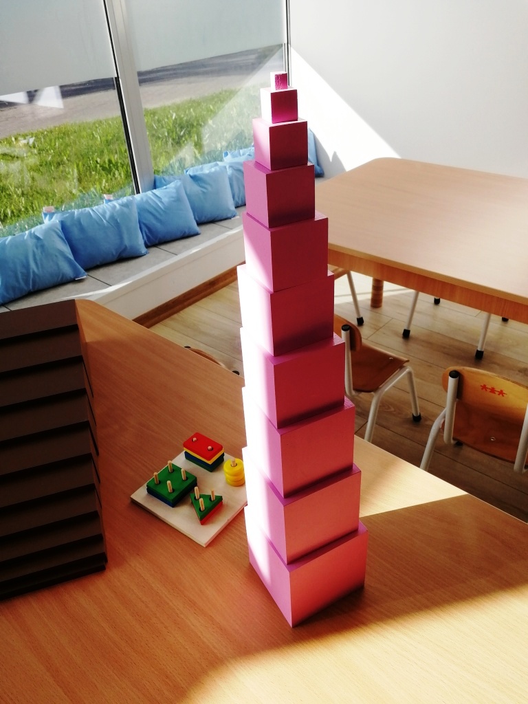 Różowa wieża z drewnianych sześcianów o różnej wielkości, ułożona w piramidę, obok drewniane zabawki edukacyjne na stole w jasnym pomieszczeniu z siedziskami przy oknie.