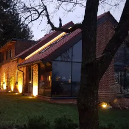Oświetlony dom z cegły z dużymi oknami w stylu nowoczesnej stodoły, podświetlony liniowym światłem LED na dachu i reflektorami w ogrodzie po zmierzchu.