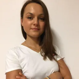 Portret uśmiechniętej młodej kobiety w białej koszulce z rękami skrzyżowanymi na tle białej ściany, widoczny delikatny złoty łańcuszek i bransoletka.